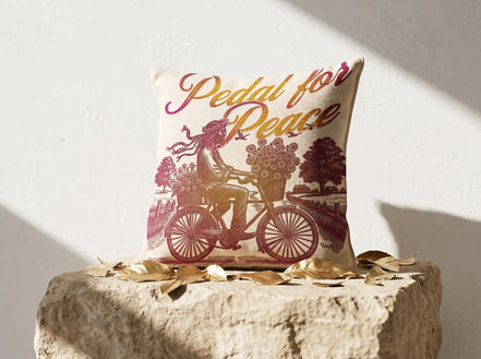 Coussin carré Essences Agape en coton écru avec illustration "Pedal for Peace" d'une personne à vélo chargée de fleurs, dégradé rose et doré.