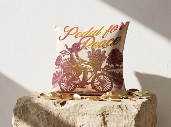 Coussin carré Essences Agape en coton écru avec illustration "Pedal for Peace" d'une personne à vélo chargée de fleurs, dégradé rose et doré.