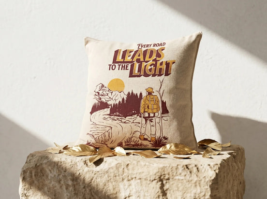 Coussin Essences Agape en coton écru avec illustration d'un randonneur regardant vers la lumière, texte 'Every Road Leads to the Light'.