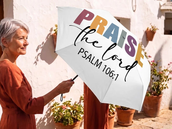 Parapluie pliant blanc Essences Agape avec inscription "Praise the Lord Psalm 106:1" en lettres multicolores. Une femme tient le parapluie ouvert.