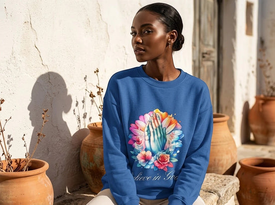 Sweatshirt bleu roi Essences Agape avec un imprimé de mains jointes en prière entourées de fleurs colorées et l'inscription "Believe in God".