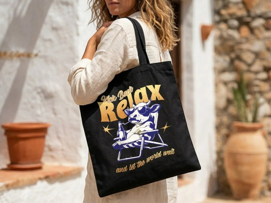 Sac fourre-tout noir Essences Agape en coton avec un imprimé humoristique de vache relaxant sur une chaise longue, lunettes de soleil et boisson.