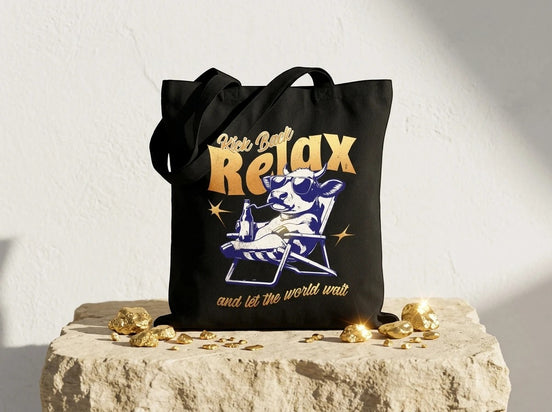 Essences Agape tote bag noir en coton avec un imprimé humoristique de vache détendue sur une chaise longue, lunettes de soleil et boisson. Message "Kick Back Relax".